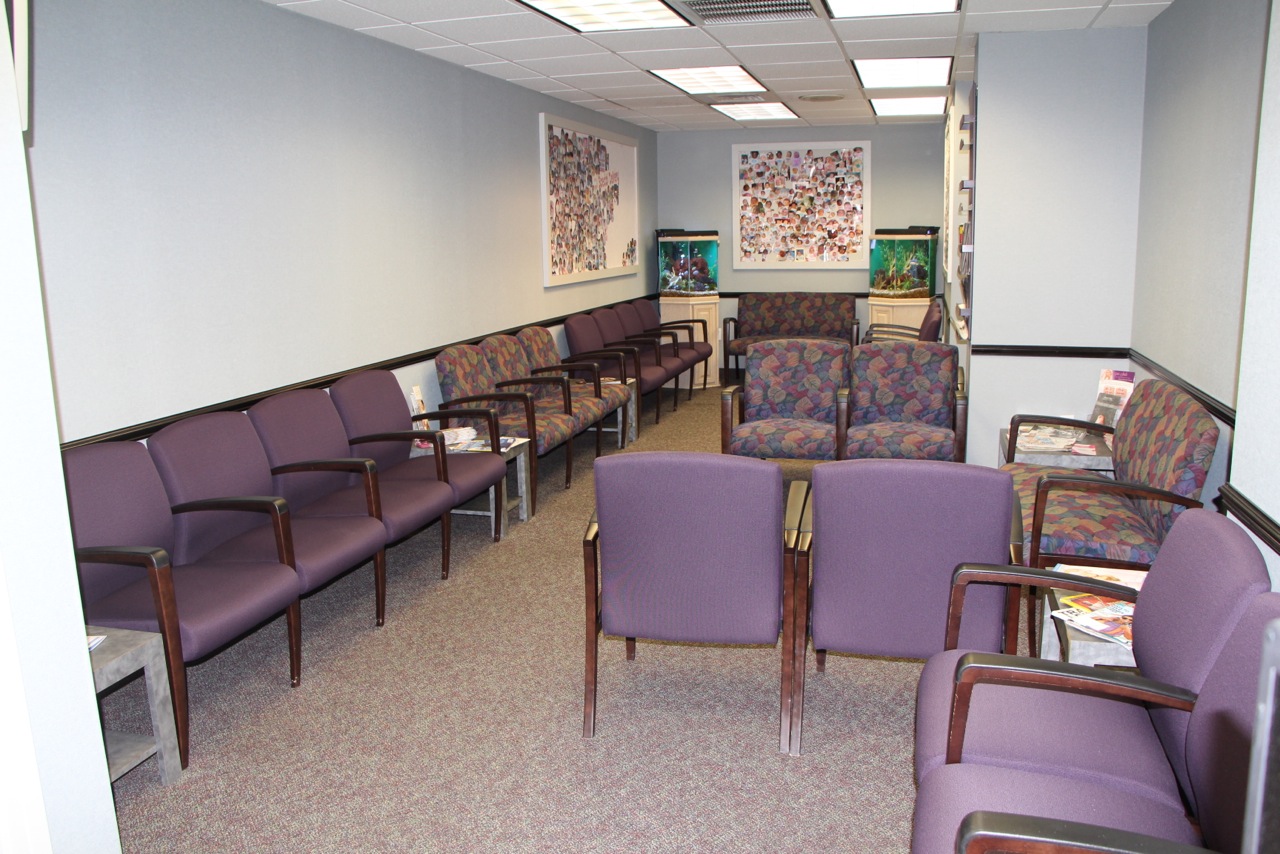 A waiting room with rows of purple chairs, small tables with magazines, two fish tanks, artwork on the walls, and fluorescent ceiling lights. The room is empty and clean.