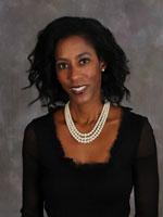 A woman with shoulder-length black hair wearing a sheer black top and a multi-strand pearl necklace poses in front of a gray mottled background, smiling slightly at the camera.
