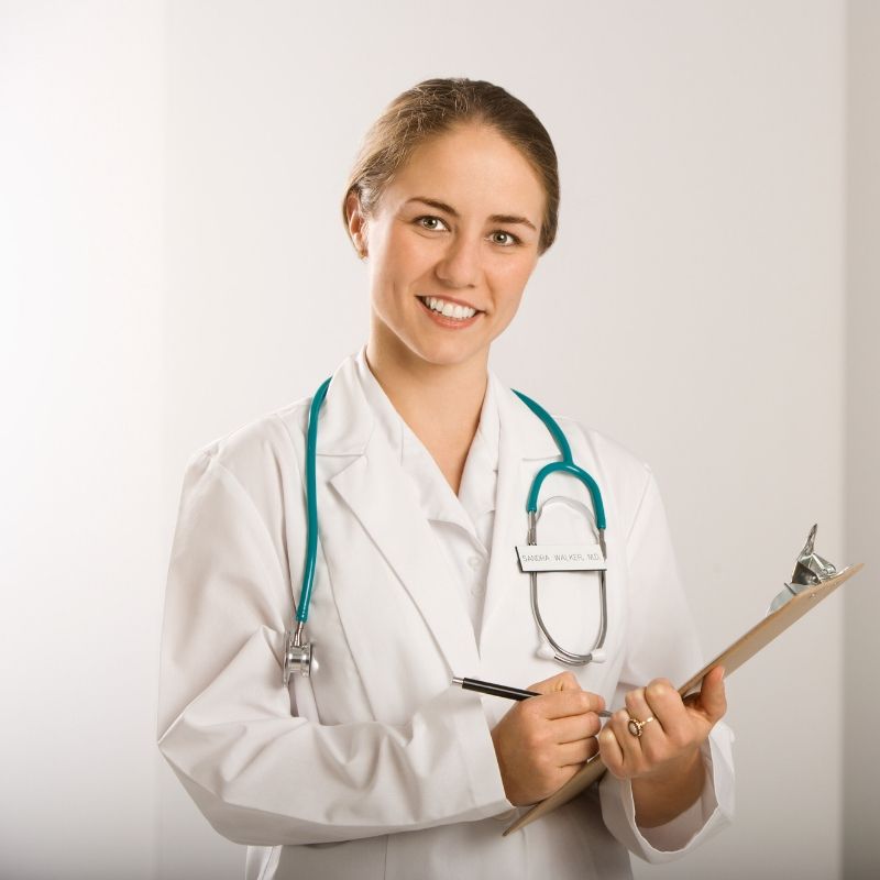 A smiling female doctor wearing a white lab coat and a teal stethoscope around her neck holds a clipboard and pen, standing in a brightly lit room.