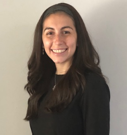 A woman with long brown hair, wearing a black top and a headband, stands smiling in front of a plain light-colored wall.