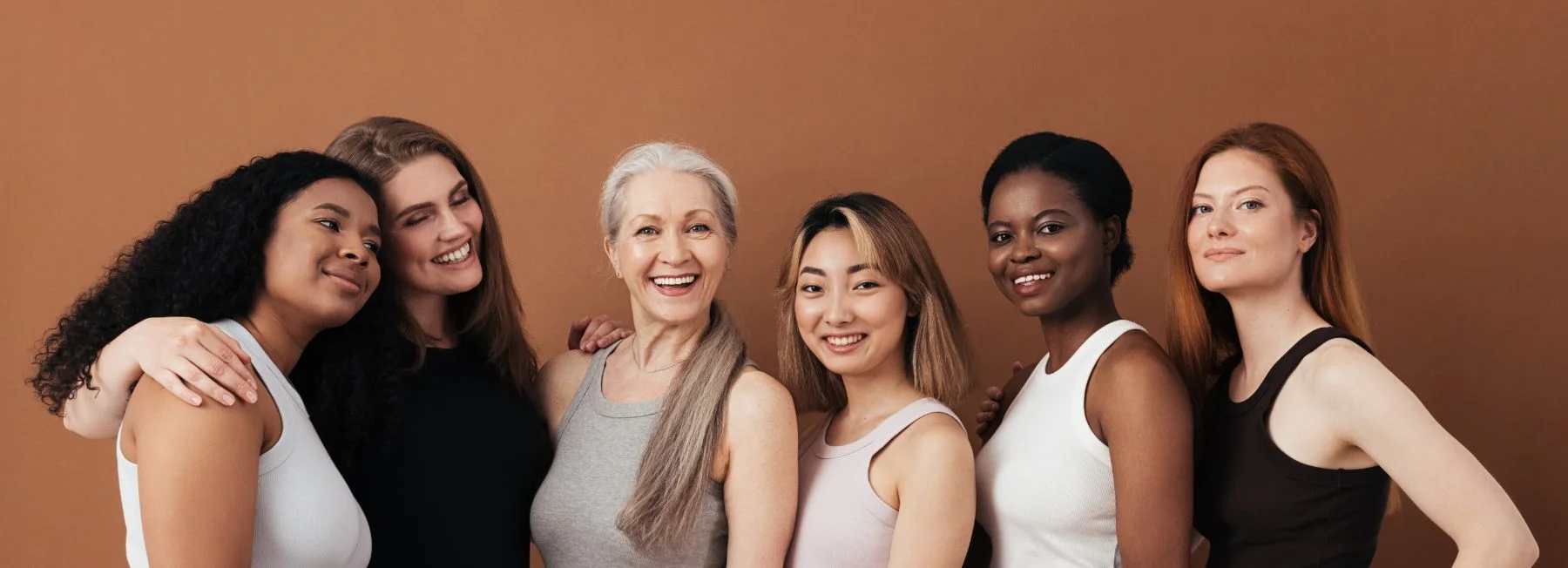 Six women of diverse ages and ethnicities stand together in front of a brown background, smiling and posing closely as a group.