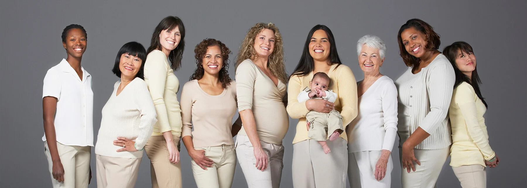A diverse group of women of different ages and ethnicities stand together, smiling at the camera. One woman holds a baby. They are dressed in neutral-colored clothing against a plain gray background.